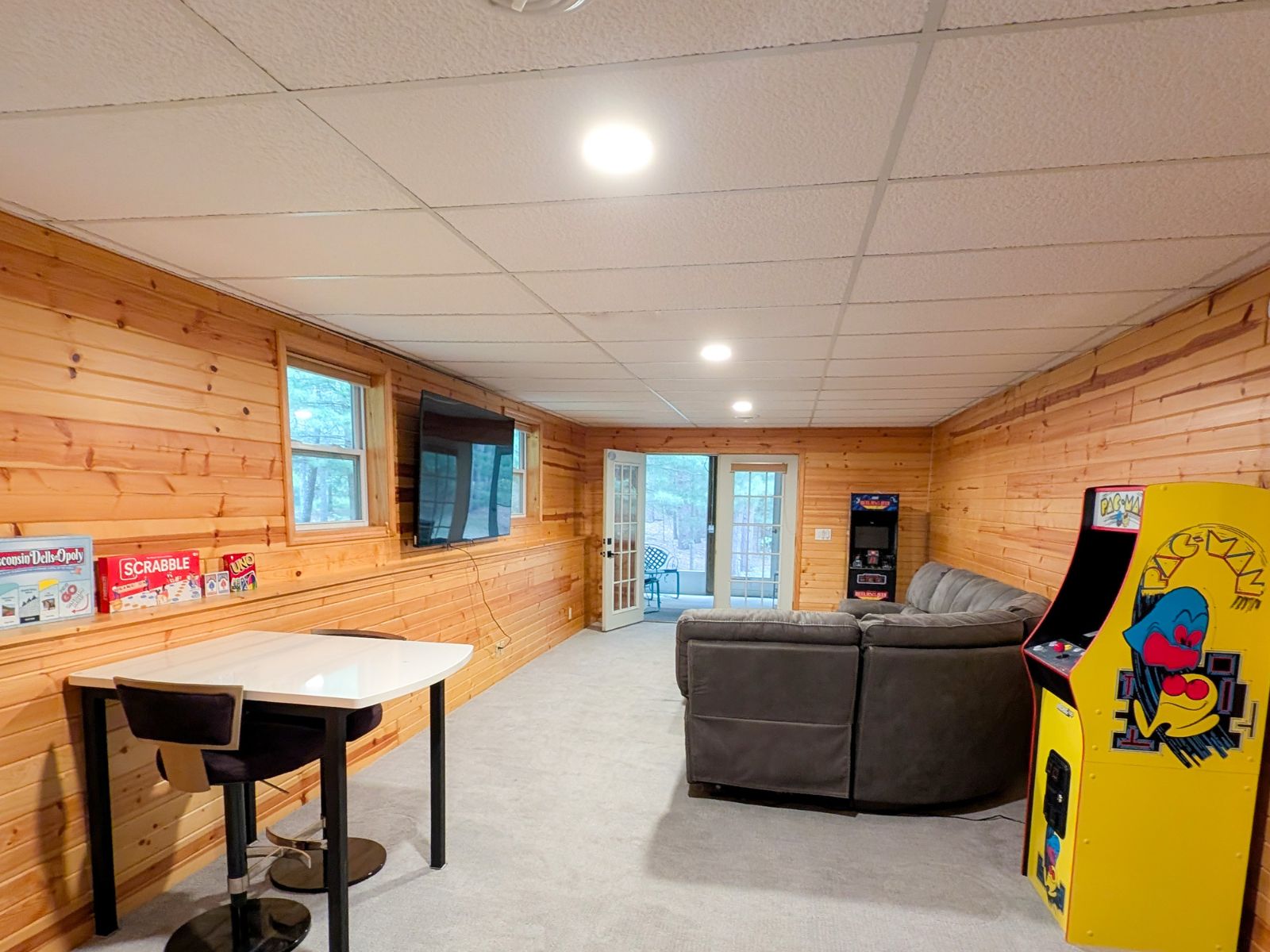 Wide view of the finished game room with arcade games, television, seating, game shelf, and porch doors